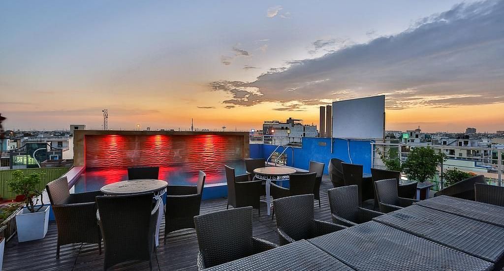 A rooftop terrace during sunset with wicker furniture, a lit pool and a view of the city skyline - TGI Apple Inn, Jaipur 