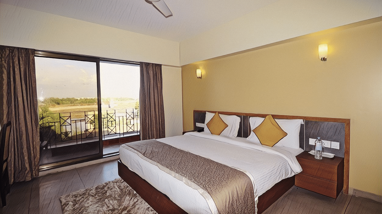 An image of a bedroom which also has a balcony from where the outside world can be seen - Kamats India