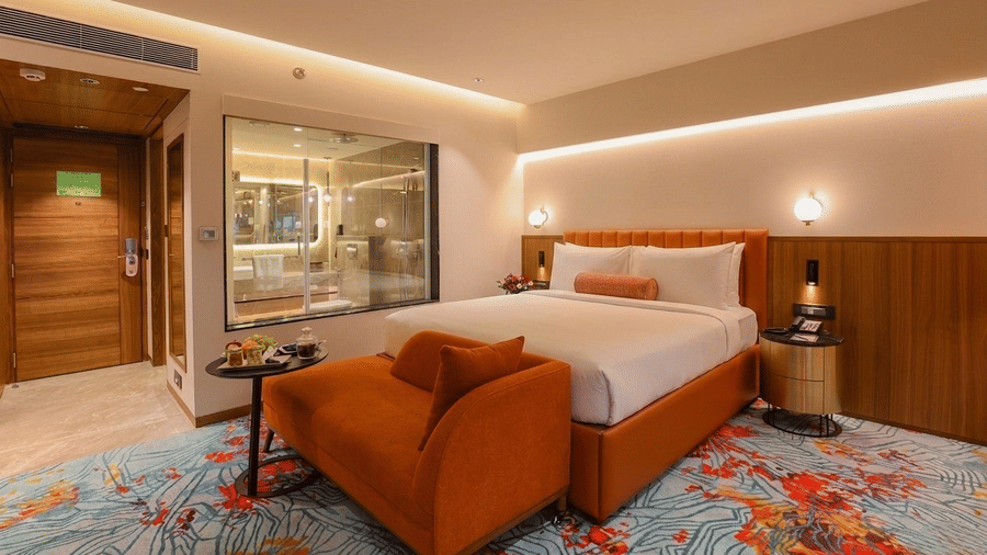 Hotel room at Pride Premier Pune with an orange bed frame, matching armchair, patterned carpet, and glass-walled bathroom.