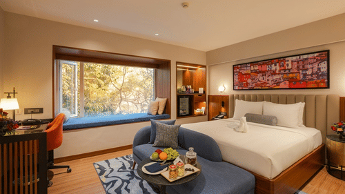 Pride Premier Pune hotel room featuring a double bed, blue sofa, window seat, work desk, and a colourful painting on the wall.