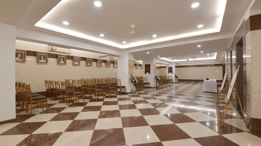 An event hall with brown and beige chequered floor featuring rows of chairs, photographs hanging high on the wall, and a few placards resting by the side.