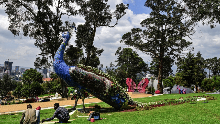 A large peacock sculpture stands in a green park where visitors relax on the grass.