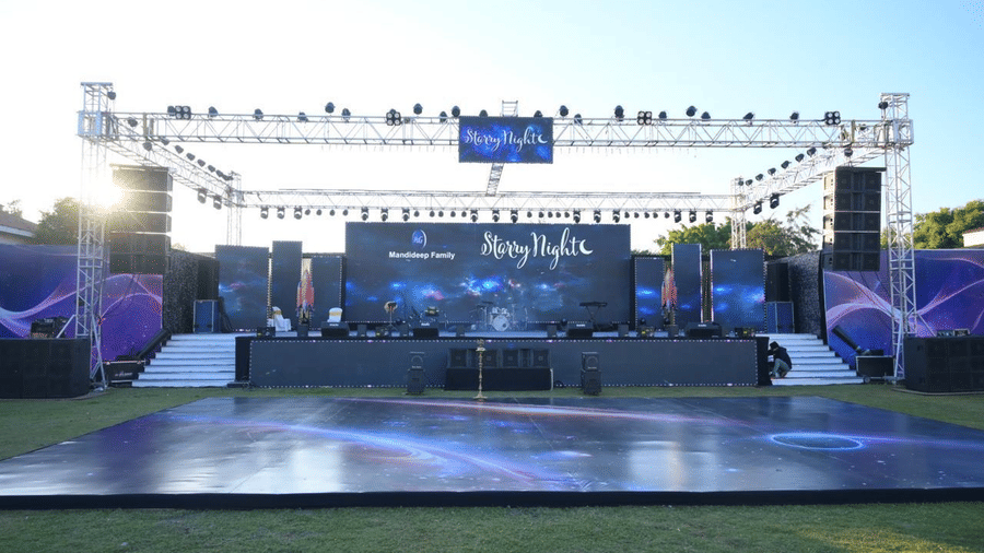 An outdoor stage setup with truss lighting, a large backdrop, and open lawn space in front.