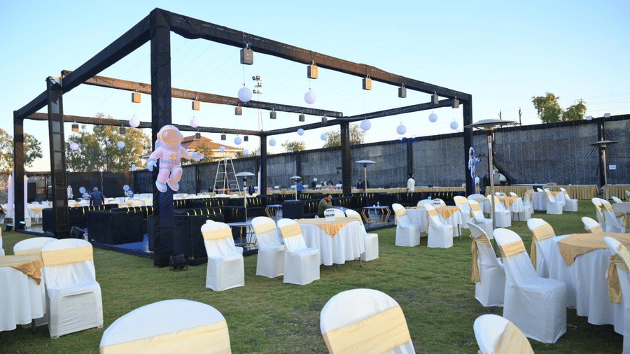 An outdoor lawn seating arrangement with round tables, covered chairs, and a metal frame structure above.