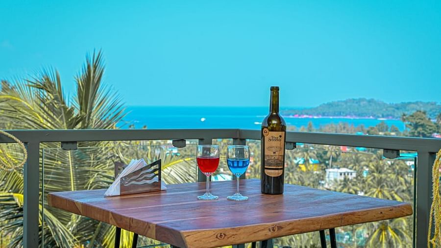A table with a bottle of wine and two glasses of colourful drinks, set outdoors on a balcony with a scenic view of the sea and palm trees under a clear blue sky - Sea Hills Hotels, Sri Vijaya Puram (Port Blair)