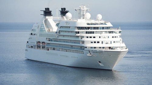 A large cruise ship sailing on calm waters, showing multiple decks, antennas, and lifeboats with the horizon visible in the distance.