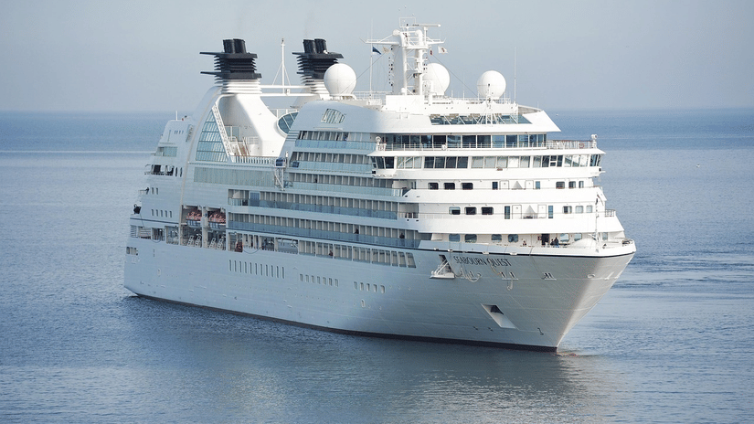 A large cruise ship sailing on calm waters, showing multiple decks, antennas, and lifeboats with the horizon visible in the distance.