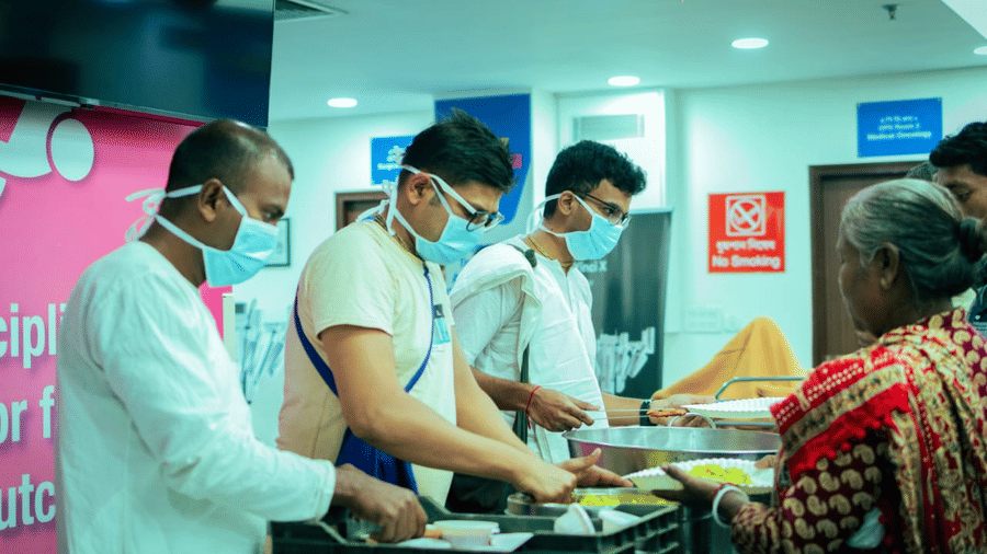 Volunteers wearing masks and aprons serve meals to a woman at HCG Hospital.