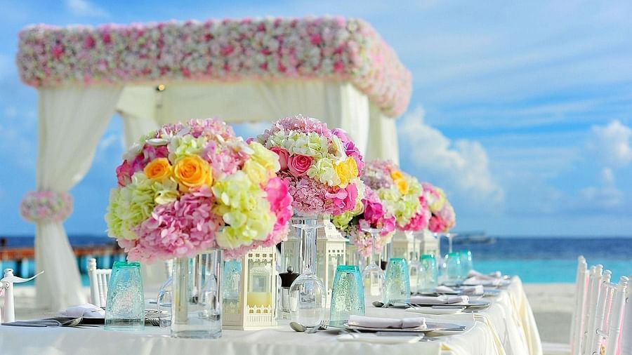 Elegant beachside wedding setup with pastel floral centrepieces, white draped canopy, and neatly arranged tableware under a bright blue sky.