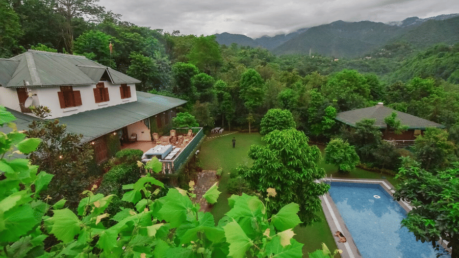 Facade of Shaheen Bagh - A Luxury Boutique Resort & Spa in Dehradun surrounded by trees with a swimming pool next to the lawn area and mountain ranges in the background.