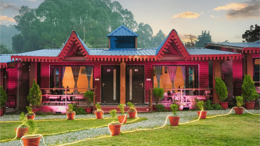 A close-up front view of two symmetrical brick cottage units under a sunset sky. The cabins feature large glass windows reflecting warm interior lights, pink accent lighting on the walls, and a decorative rope light border along the garden path in the fore
