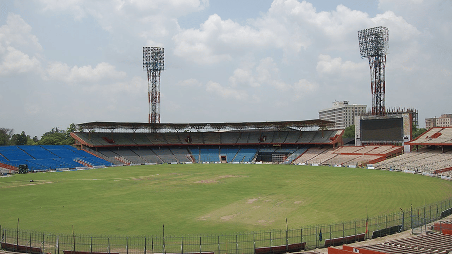 Cricket ground under clear blue sky with tall floodlight towers, practice area, and tiered stands surrounding the oval.