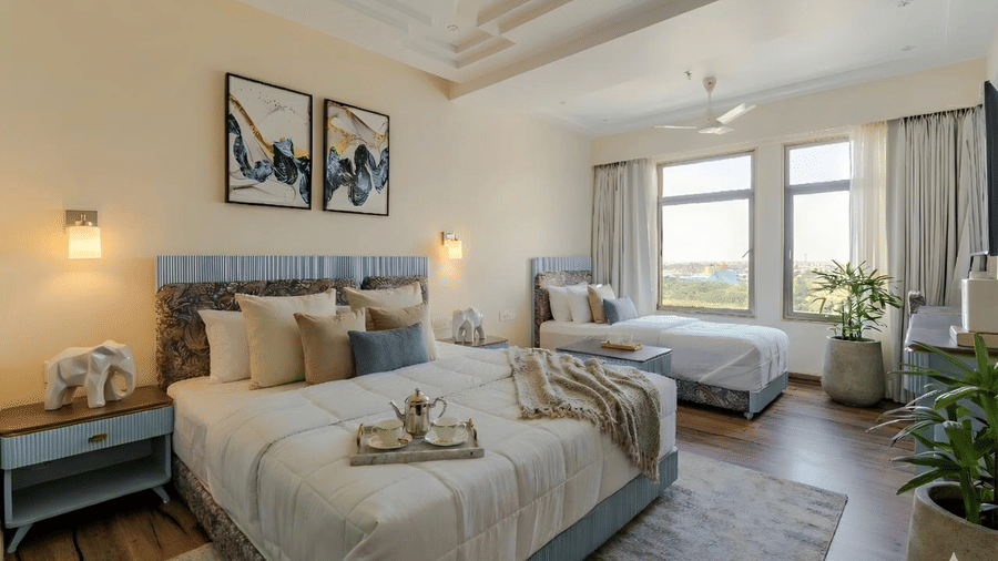 A wide-angle shot of the Deluxe Room featuring a double bed with a lot of cushions along with a single bed beside it, with ample of natural light coming from the window illuminating the entire room at Essentia Luxury Hotel and Convention, Nagpur.