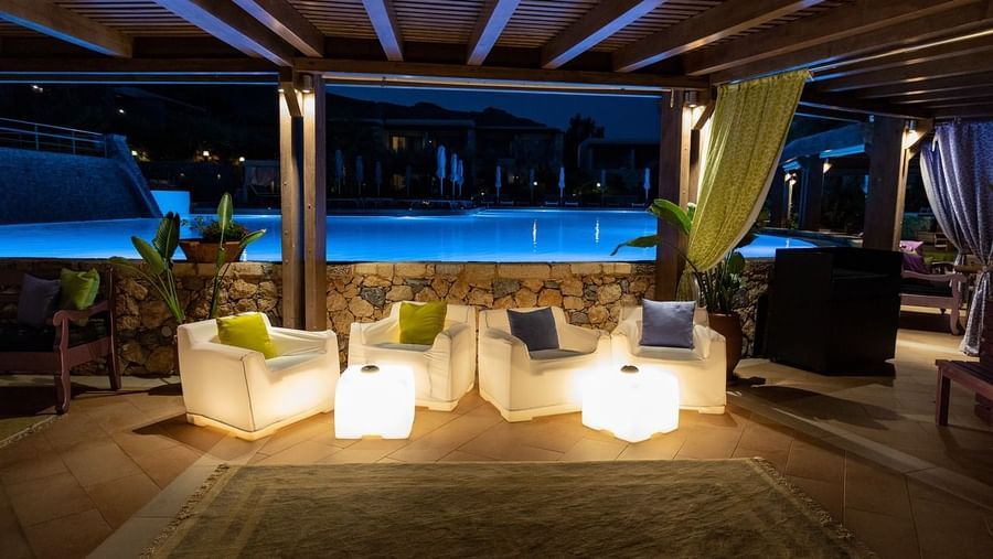 A stylish outdoor lounge area with white illuminated cube seats and a wooden pergola by a serene pool at dusk.