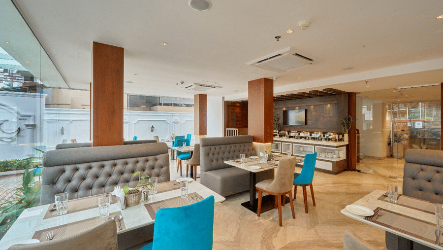 Wide view of the restaurant at QWhite Crescent Luxury Hotel by Dimora Kochi with teal chairs, cushioned booths, and warm ambient lighting.