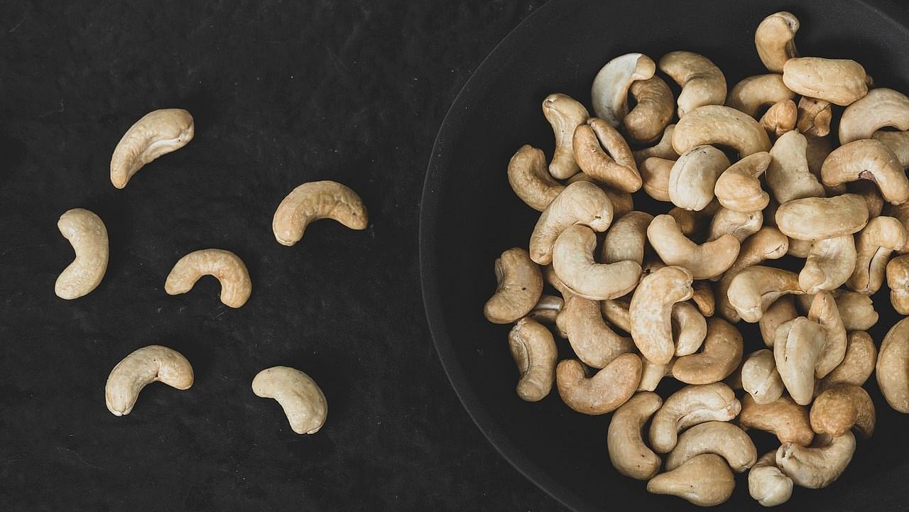 An image of a black bowl filled with cashews, with six cashews placed on one side of the bowl on a black surface.