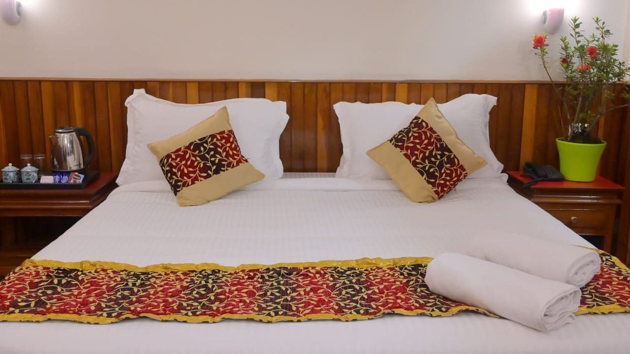 Neatly made double bed with white linens, two patterned cushions, a colourful bed runner, rolled towels, and bedside tables with a kettle, cups, and a green potted plant - Sumitel Chiminda, Pelling.