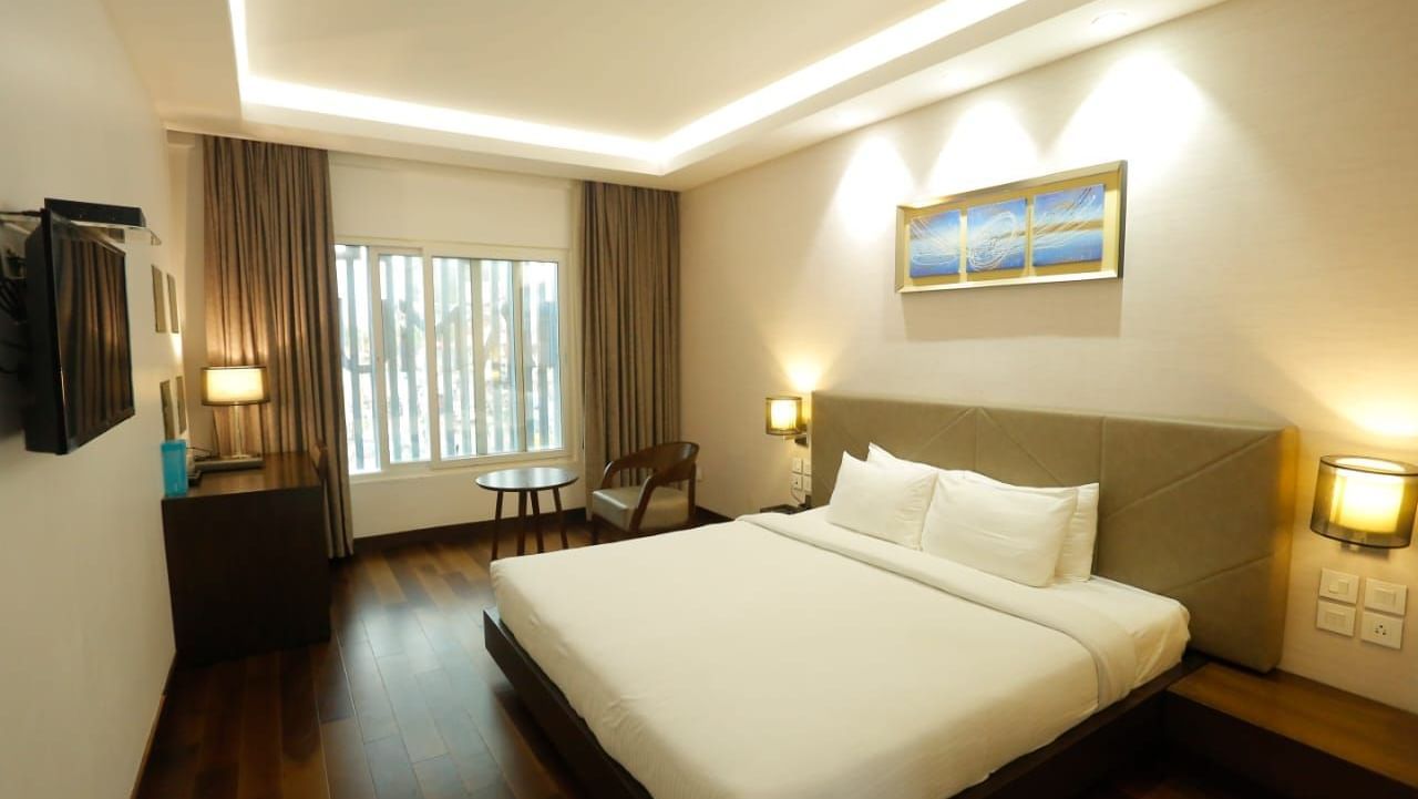 Spacious room with a large double bed, bedside lamps, wall art above the headboard, and a window with curtains pulled to the side at The Hamuse Hotel, Kodaikanal.