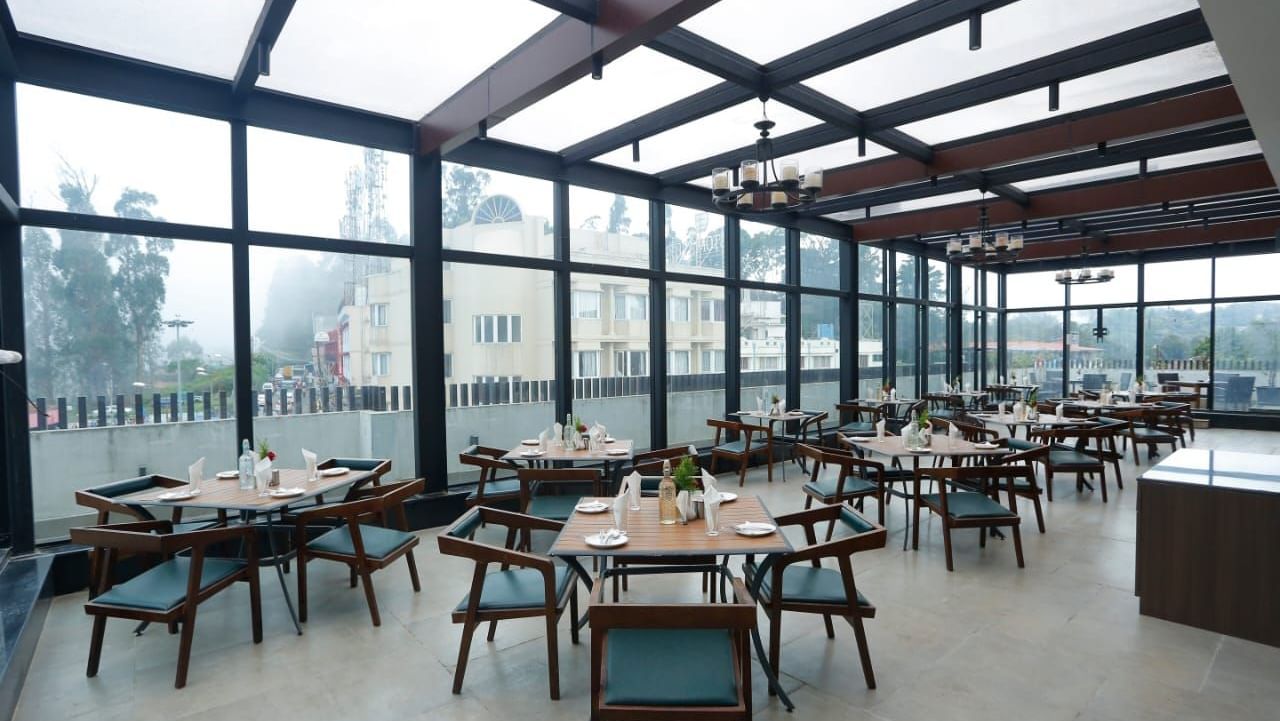 A rooftop dining area with glass walls and ceiling, offering stunning views and arranged tables and chairs for guests at The Hamuse Hotel, Kodaikanal.