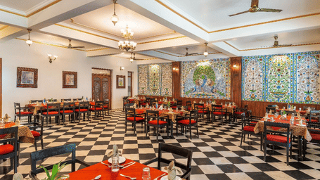 An interior view of a dining hall showing rows of tables with orange cloths arranged on a checkered floor tile pattern at Amantra Shilpi Resort, Udaipur.