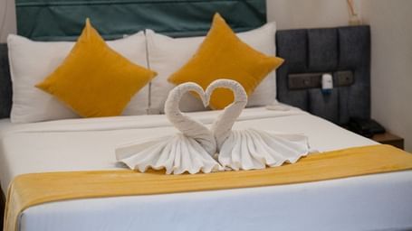 A room with a double bed, yellow pillows, and neatly arranged towels shaped into a heart, creating a romantic ambience at Hotel Kyzen Smartstays – Smart, Reliable, Kyzen