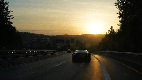 A car driving on a highway during sunset, with the sun low on the horizon and trees on both sides.