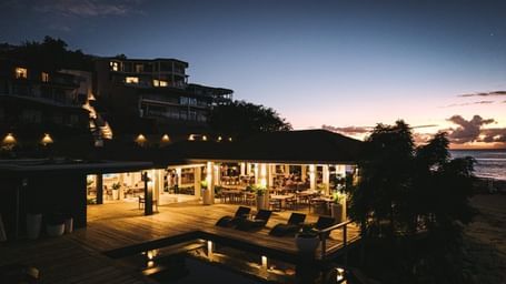 A coastal resort at dusk, with illuminated buildings, an outdoor patio, and a pool, under a twilight sky.