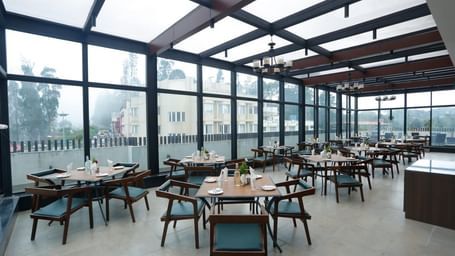 A rooftop dining area with glass walls and ceiling, offering stunning views and arranged tables and chairs for guests at The Hamuse Hotel, Kodaikanal.