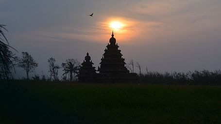 silhouette of a architectural figure with two pointed roofs in a setting of trees with the sun in the distance 
