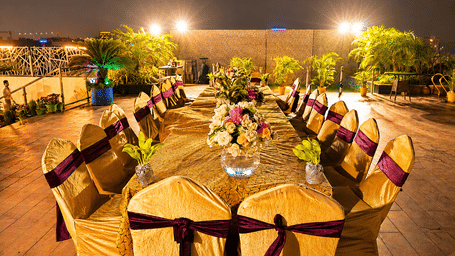Outdoor poolside deck set up at night with a long banquet table, gold chair covers with purple sashes and floral centrepieces along the table.