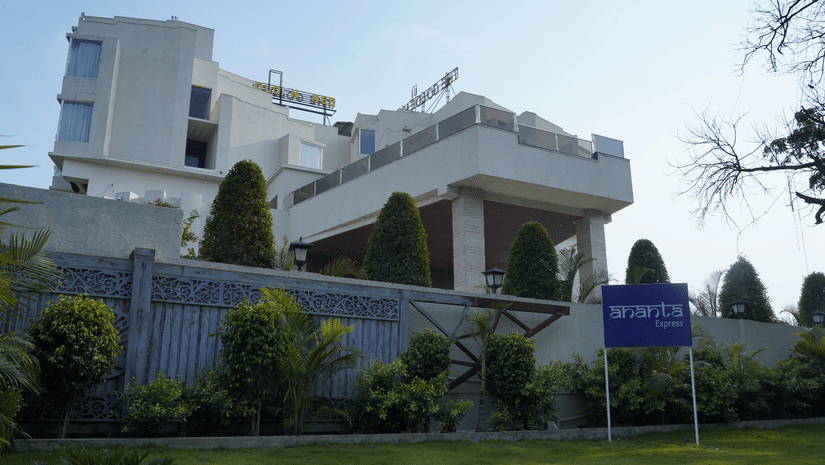 An exterior view of the hotel building behind a green lawn, trees, and an entry gate at Ananta Express, Jaunpur.