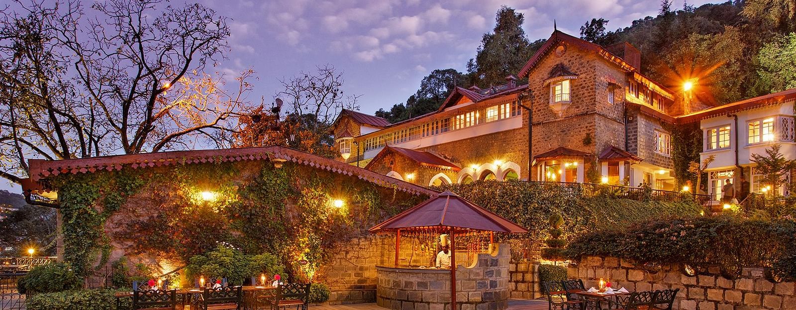 A beautiful outdoor dining area at the naini retreat, nainital. There are tables and chairs set up on a stone patio, with a fireplace in the center. The building is lit up at dusk, creating a warm atmosphere. 