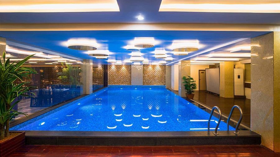 An indoor swimming pool at Mansha Hotel Regency with blue water and a light blue ceiling, surrounded by pillars and lit by overhead lights.