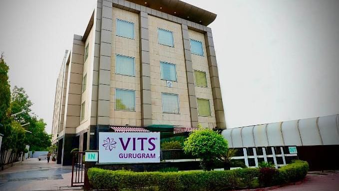 A low-angle exterior shot of a multi-storey building with a sign that reads VITS GURUGRAM, surrounded by greenery