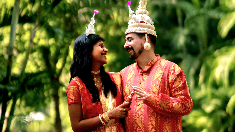A bride and groom in traditional Bengali wedding attire posing together in a lush green garden at Vedic Village Spa Resort with tropical trees visible in the background.