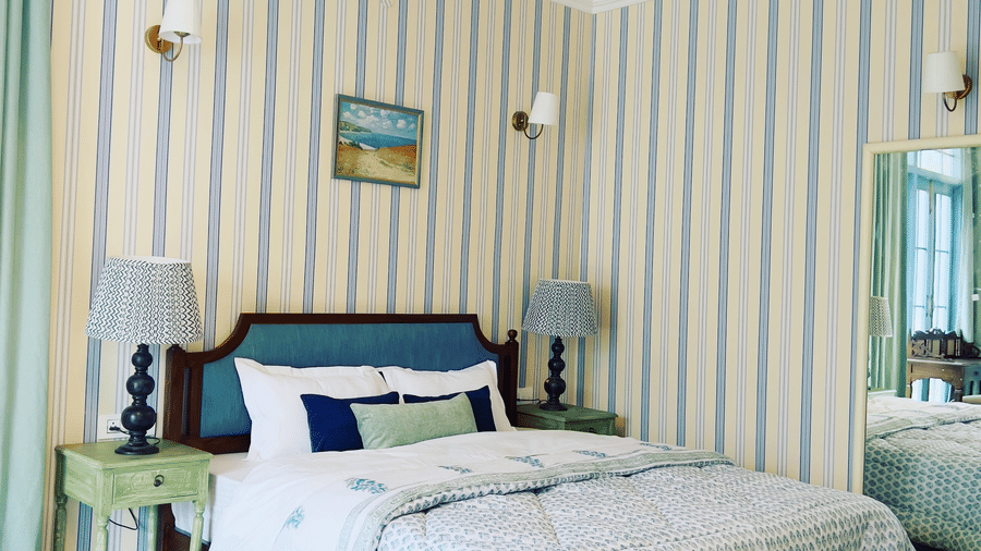 A room features vertical blue and yellow striped wallpaper, a large bed with a teal headboard, mint-green bedside tables, and matching table lamps at Faraway Cottages, one of the best places to stay in auli.