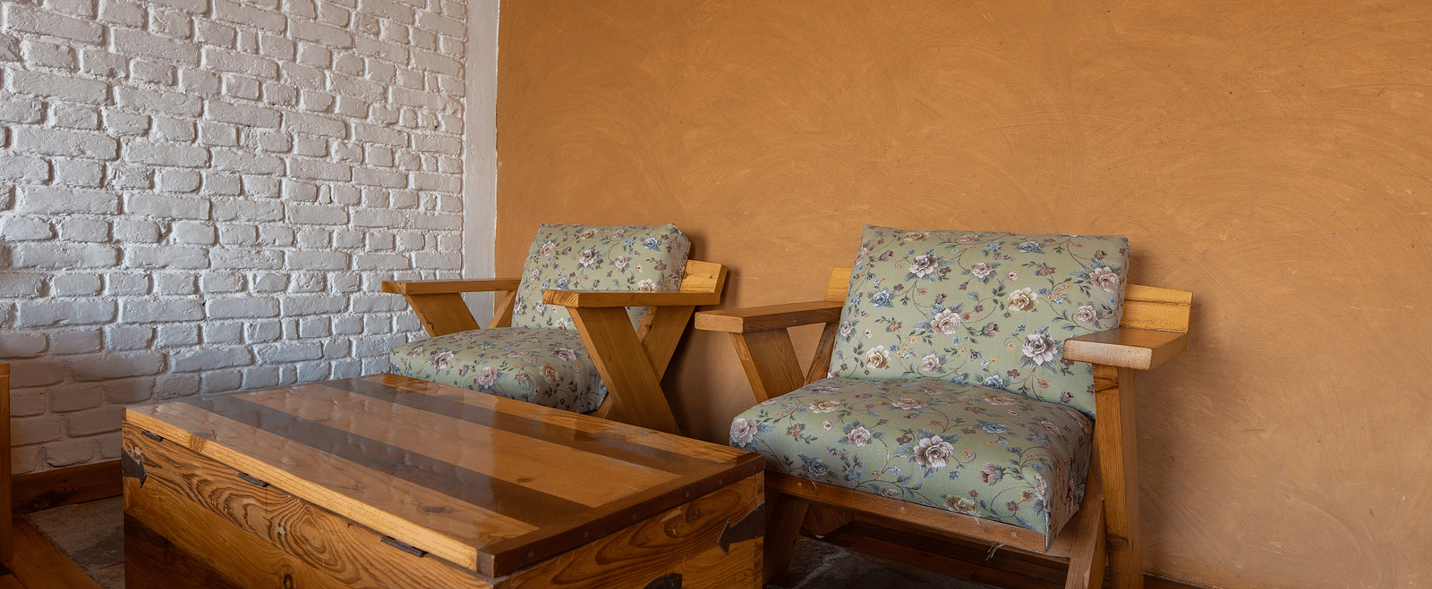 Wooden chairs with floral cushions and a chest table against a white brick wall. - Blusalzz Homes The Himalayan Bungalow, Almora