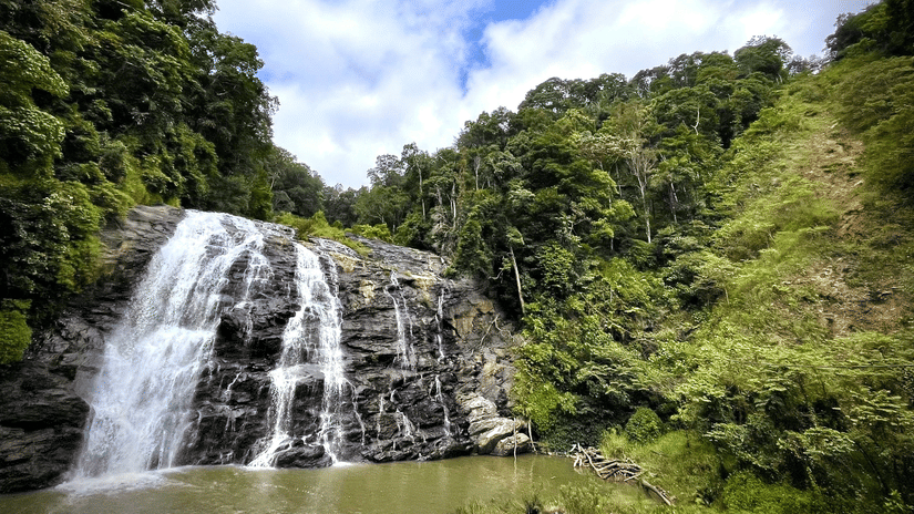 A waterfall flowing over a rocky cliff surrounded by trees and green vegetation under a partly cloudy sky.