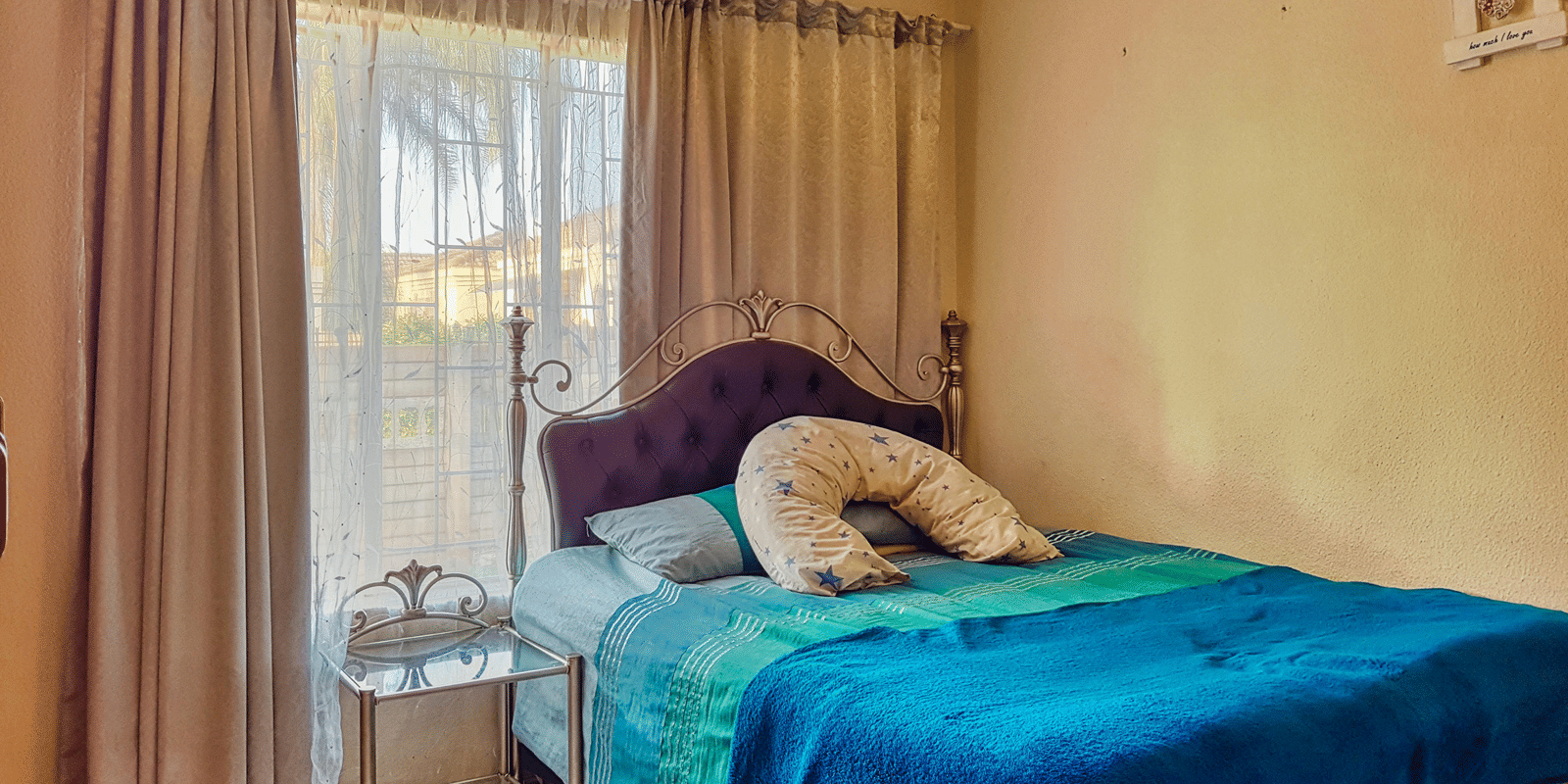 A bedroom featuring a double bed with blue sheets spread neatly over it with beige curtains covering the window behind it.