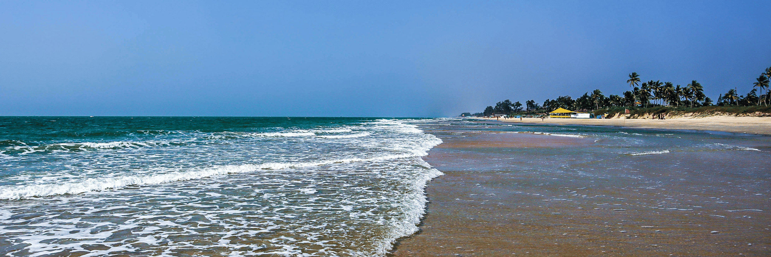 A view of Varca Beach, one of the most popular beaches in South Goa, with the foamy waves crashing on the shoreline with trees in the distance and blue sky above.