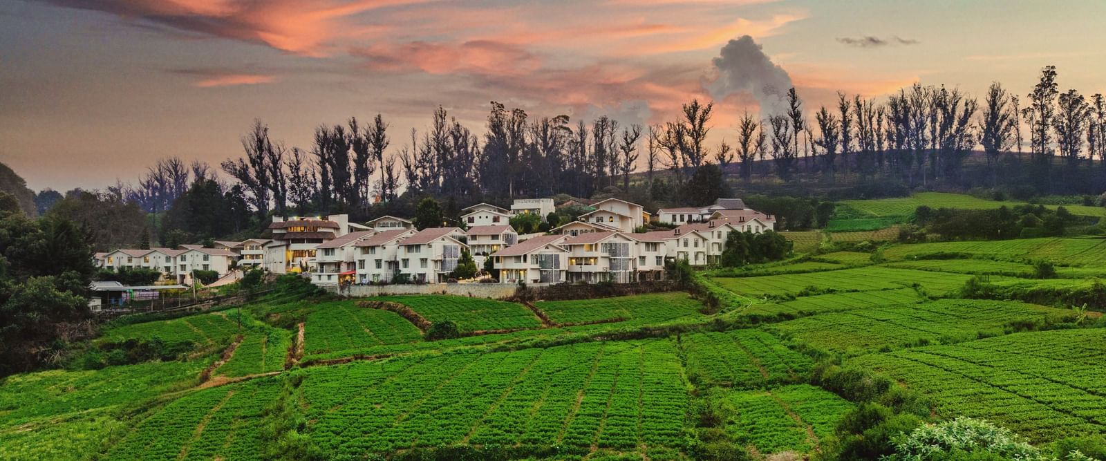 Lush green fields with a backdrop of white cottages nestled under a vibrant sunset sky in a hilly landscape.