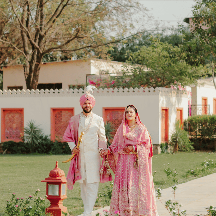 A bride and groom dressed in traditional pink and white wedding attire walk together along a pathway next to a green lawn at Umaid Palace.