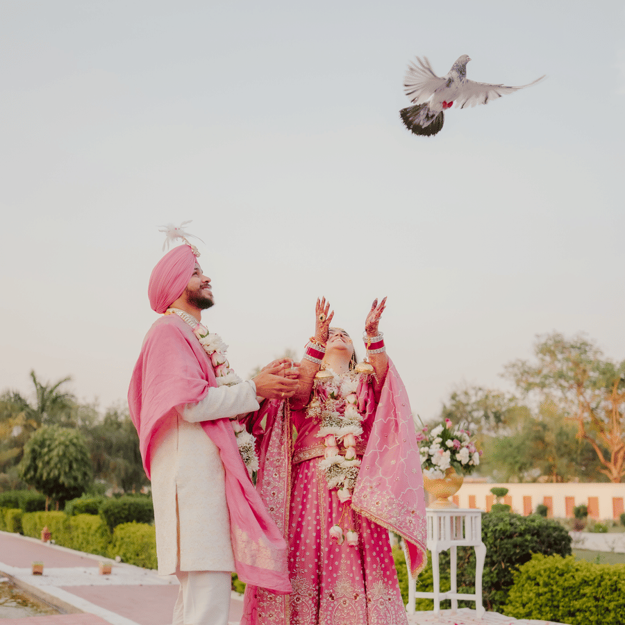 A bride and groom stand together on a path covered in pink flower petals while releasing a pigeon into the air during their wedding at Umaid Palace.