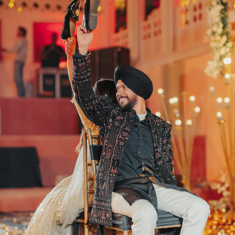 Groom Jaswinder holding shoe while seated back?to?back with bride Ritika during playful wedding game at Umaid Palace celebration.