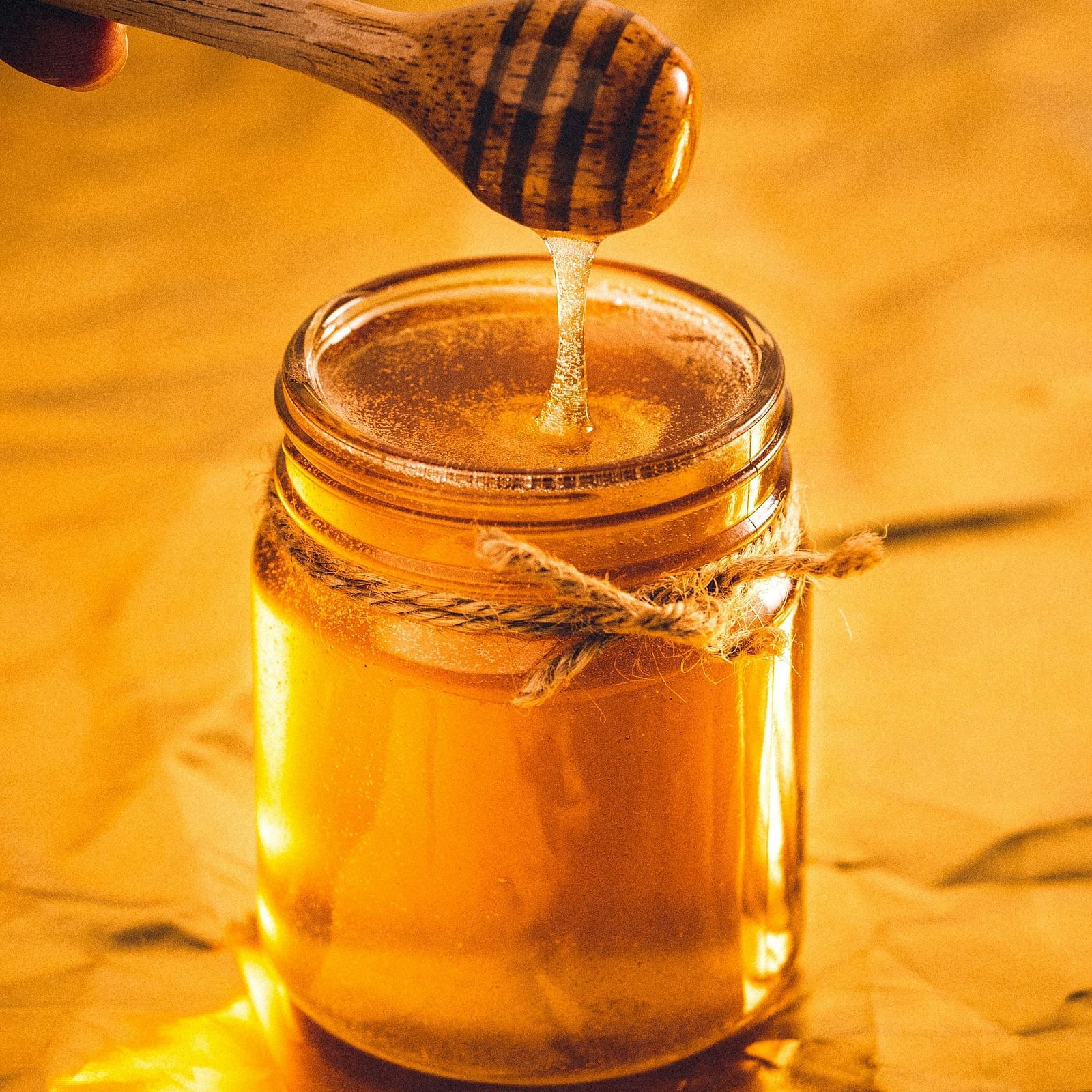 Golden honey dripping from a wooden dipper into a glass jar