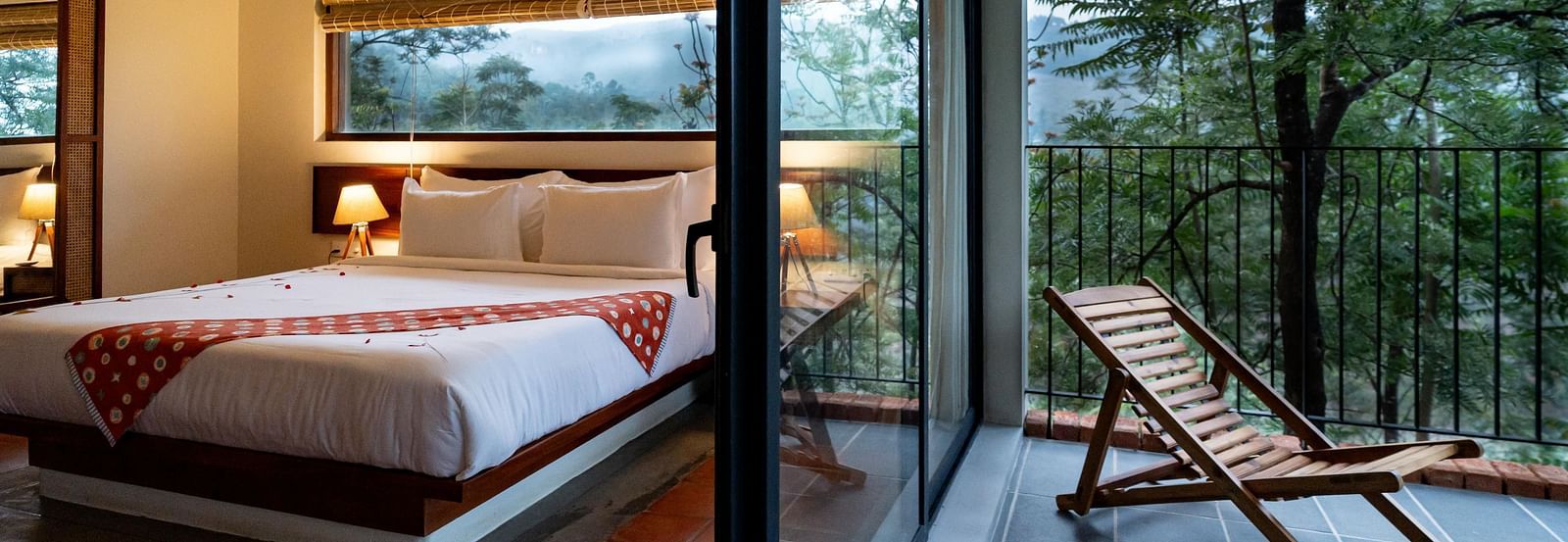 A spacious room at Vaasta Vagamon with a comfortable bed, leading to a private balcony with a wooden lounge chair and scenic views.