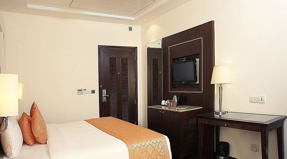 Executive Room with a double bed, bedside table with a lamp on top, and a wall-mounted TV & wooden table with lamp at Inde Signature Tower, Gurgaon