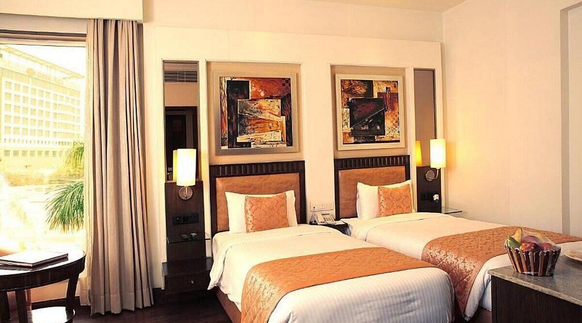 The Premium Twin Room offers twin beds with neatly arranged pillows, wooden bedside tables with lamps, a cushioned seating area with a wooden table, and large side windows for natural light at Inde Signature Tower, Gurgaon