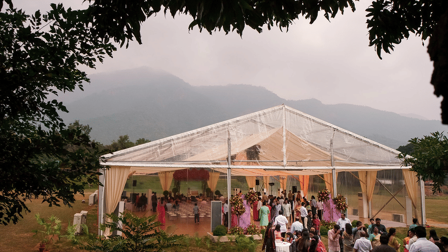 An outdoor event with guests gathered near a clear marquee tent, surrounded by nature and mountains in the background, creating a scenic and festive atmosphere at Black Thunder, Coimbatore