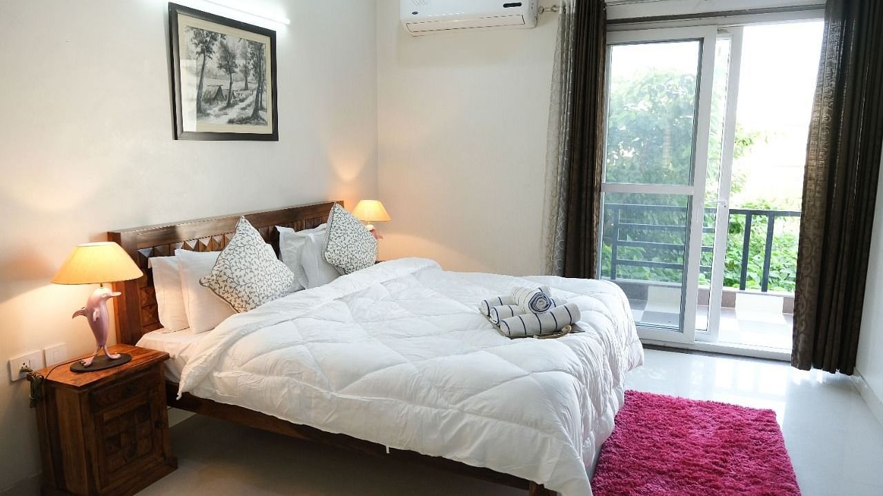 An image of a bedroom featuring a bed, a bedside table with lamp, anda a glass door leading to the balcony - LIme Tree 3BHK Serviced Apartments Near Medanta HospitalAn image of a bedroom featuring a bed, a bedside table with lamp, anda a glass door leading